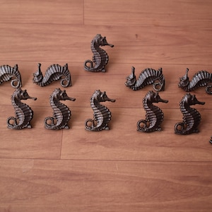 May include: Bronze seahorse-shaped cabinet pulls and screws are arranged on a wooden surface. The seahorse pulls have textured surfaces and are in various poses. Suitable for nautical or coastal decor, these pulls are decorative.