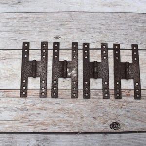 May include: A set of four rectangular metal hinges with a dark, weathered, and rusted finish. Each hinge features a central barrel and two parallel vertical plates, each containing six small circular screw holes for mounting purposes.