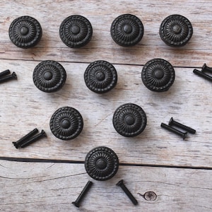 Puede incluir: Diez tiradores de armario redondos negros con un diseño floral detallado. Los tiradores están acompañados de varios tornillos negros, todos dispuestos sobre una superficie clara con vetas de madera. Ideales para la restauración de muebles.