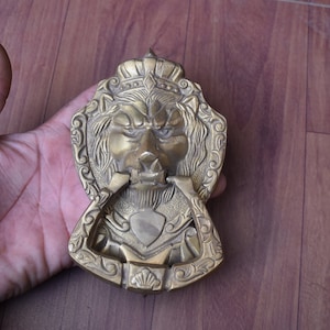 Könnte beinhalten: Ein goldfarbener Türklopfer in Form eines Löwenkopfes mit filigranen Details. Der Türklopfer ist in Form eines Löwenkopfes mit einer Krone auf dem Kopf. Der Löwenmaul ist offen und hat einen Ring zum Klopfen.