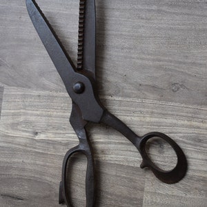 May include: A pair of vintage black metal shears with a serrated blade and a wooden handle. The shears are lying on a wooden surface.