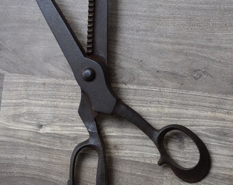 May include: A pair of vintage black metal shears with a serrated blade and a wooden handle. The shears are lying on a wooden surface.