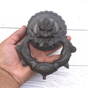 Peut inclure: Un heurtoir de porte orné en bronze foncé, avec un motif de visage détaillé et une poignée en forme d'anneau. Le heurtoir est tenu dans une main sur un fond en bois blanc. Le design présente un visage avec un motif en forme d'éclat de soleil.