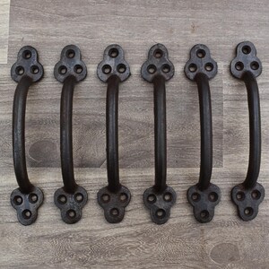 Peut inclure: Six poignées en fonte noire avec un design incurvé. Chaque poignée a une base décorative avec trois trous et une poignée incurvée. Les poignées sont disposées en rangée sur une surface en bois.