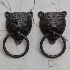 Puede incluir: Dos llamadores de puerta de hierro fundido negro en forma de cabeza de oso con anillos redondos. Las cabezas de oso tienen detalles y están montadas sobre una superficie de mármol blanco.