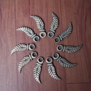 Könnte beinhalten: Zehn silberfarbene, blattförmige Metallanhänger mit einem Loch oben. Die Anhänger sind in einem Kreis auf einer Holzoberfläche angeordnet.