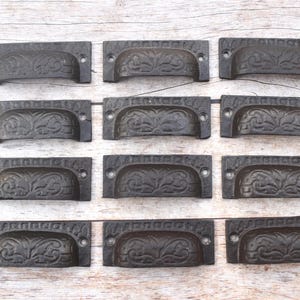 Pode incluir: Um conjunto de doze puxadores de gaveta em metal castanho escuro dispostos numa grelha. Cada puxador retangular apresenta uma secção curva com um padrão floral em relevo e dois orifícios circulares para parafusos nas abas de montagem.