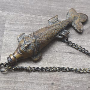 Könnte beinhalten: Bronze-farbener Flaschenverschluss in Form eines Fisches mit Kette. Der Fisch hat detaillierte Schuppen, Flossen und ein Gesicht. Die Kette besteht aus kleinen, ineinandergreifenden Gliedern. Der Verschluss ist wahrscheinlich für eine Weinflasche.