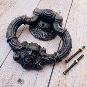 Puede incluir: Un llamador de puerta de hierro fundido negro con un diseño detallado que presenta una cara y un trabajo de desplazamiento intrincado. El llamador está unido a una puerta de madera con dos tornillos.