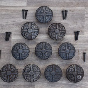 May include: A collection of ten dark gray, round cabinet knobs with an ornate, symmetrical design. Each knob is accompanied by a set of black screws. The knobs are arranged on a light gray wood-grain surface.
