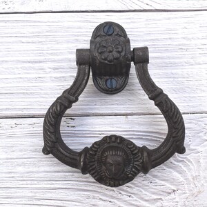 May include: A dark bronze door knocker. The oval backplate has a floral design and two screws. The knocker has a curved handle with a detailed rope-like texture and a face design at the bottom. The door knocker is made of metal.