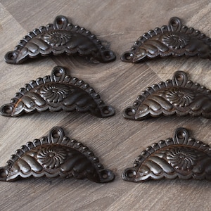 May include: Six antique-style, dark brown, cast iron drawer pulls with a decorative floral design. Each pull is shaped like a half-circle with a scalloped edge.