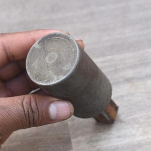 Op de afbeelding: Een cilindrisch metalen object met een houten handvat. Het metalen deel is dof zilverkleurig met concentrische cirkelvormige patronen aan de bovenkant. De handgreep is donkerbruin. Het object wordt in een hand gehouden.
