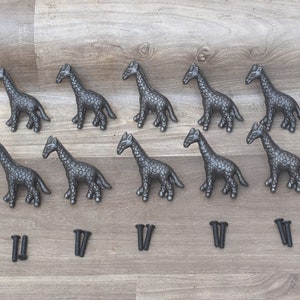 Puede incluir: Diez tiradores de cajón de metal negro en forma de jirafa con tornillos. Los tiradores están en una fila sobre una superficie de madera.