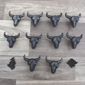 Op de afbeelding: Een verzameling van tien zwarte gietijzeren kastknoppen in de vorm van een stierschedel. Elke schedel heeft gedetailleerde horens en gelaatstrekken. De knoppen zijn gerangschikt op een lichtgrijze houten ondergrond, met schroeven.