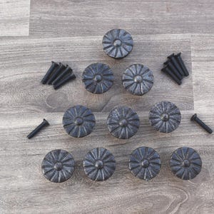 Può includere: Un set di dieci tiretti per cassetti in metallo nero con un design floreale decorativo. I tiretti sono disposti a forma di piramide su una superficie in legno.