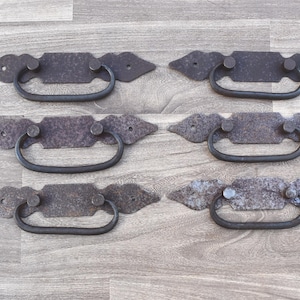 Puede incluir: Seis tiradores de cajón de hierro de estilo antiguo con un acabado rústico. Cada tirador tiene una placa rectangular con un diseño decorativo y una manija curva.