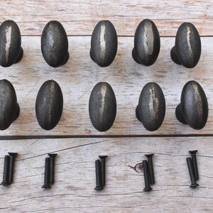 Puede incluir: Diez pomos de armario ovalados de color gris oscuro con una raya plateada vertical en el centro. Los pomos están dispuestos en dos filas sobre una superficie de madera marrón claro, con tornillos negros debajo.