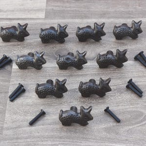 Op de afbeelding: Een verzameling van tien donker bronzen kastknoppen in de vorm van konijnen, samen met verschillende zwarte schroeven. De knoppen liggen op een lichtgrijze houten ondergrond.
