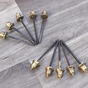 May include: A collection of antique-style furniture tacks. Each tack features a dark, slender shaft topped with a decorative, aged brass head. The tacks are arranged on a gray wood-grain surface.