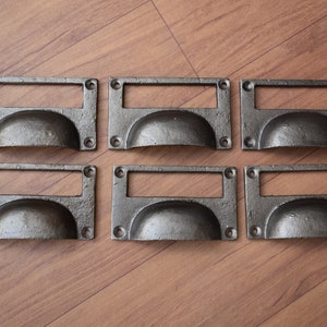 Puede incluir: Seis tiradores de cajón de estilo antiguo con un acabado oscuro y envejecido. Cada tirador presenta una placa trasera rectangular con un asa curvada y un portaetiquetas. El herraje es de metal y tiene una estética vintage.