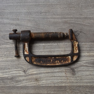 May include: A rusty metal C-clamp with a threaded screw and a handle. The clamp is on a wood surface.