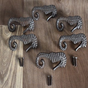 Könnte beinhalten: Acht silberfarbene, metallene Schubladengriffe in Form von Seepferdchen mit detailliertem Design. Die Griffe sind in einem Kreis auf einer Holzoberfläche angeordnet, mit verstreuten Schrauben.