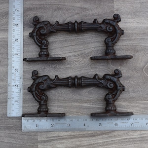 May include: Two black cast iron door handles with ornate scrollwork and a flat base. The handles are approximately 12.7 cm long and 5 cm wide.