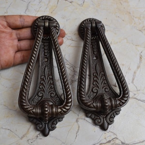 May include: Two ornate, dark brown cast iron door knockers with intricate floral designs. Each knocker has a rope-like border and a ring handle.