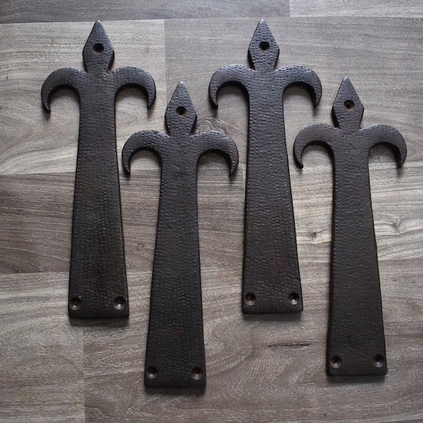 May include: Five black metal decorative hinges with a fleur-de-lis design. The hinges are arranged in a row on a wood surface.