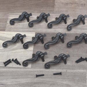 Op de afbeelding: Twaalf zwarte gietijzeren trekknoppen in de vorm van een zeepaardje met een gedetailleerd ontwerp. De knoppen zijn gerangschikt in drie rijen van vier. Er zijn ook verschillende zwarte schroeven inbegrepen.