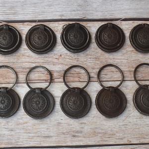 Può includere: Dieci pomelli per cassetti antichi in bronzo con un design circolare e una maniglia ad anello. I pomelli sono disposti in due file da cinque, mostrando i loro dettagli intricati e l'aspetto invecchiato. Ideale per il restauro di mobili.