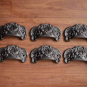 Puede incluir: Seis tiradores de cajón de hierro fundido negro con un diseño ornamentado que presenta una lira y motivos florales. Los tiradores están dispuestos en dos filas de tres sobre una superficie de madera.