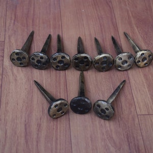 Puede incluir: Un conjunto de 13 clavos de metal negro y dorado de estilo antiguo con cabezas redondas decorativas. Los clavos tienen un acabado martillado y están dispuestos en dos filas sobre una superficie de madera.