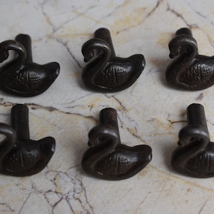 Op de afbeelding: Zes donkerbruine zwanenvormige lade- of kastknoppen. Elke zwaan heeft een gebogen nek en een gedetailleerd lichaam. De knoppen zijn gerangschikt op een lichtgekleurd, marmerachtig oppervlak.