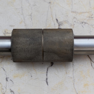 Puede incluir: Un rodillo de metal plateado con una superficie texturizada. El rodillo está hecho de dos secciones que están conectadas por una varilla de metal.