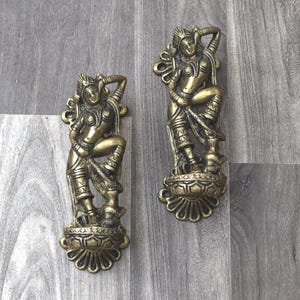May include: Two gold-toned metal door handles with a decorative design featuring a woman in a dancing pose. The handles are mounted on a wooden surface.