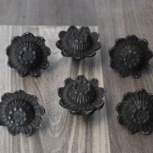 Può includere: Sei pomelli per armadi a forma di fiore, grigio scuro. Ogni pomello presenta un design floreale dettagliato con un'area centrale strutturata e bordi a forma di petalo. I pomelli sono disposti su una superficie di legno.