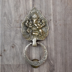 May include: A brass door knocker shaped like a leaf with a detailed depiction of the Hindu deity Ganesha. The knocker has a circular ring for pulling.