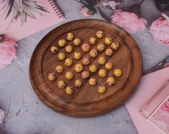 Puede incluir: Un tablero de juego de madera con una bandeja circular de madera y 30 canicas amarillas con motas moradas y azules.
