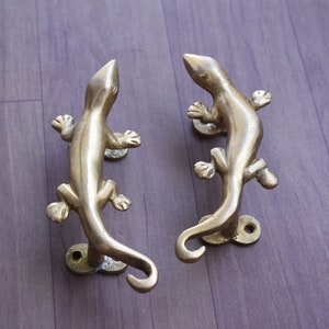 May include: Two brass lizard-shaped door handles. The handles are detailed with legs, a tail, and a head. The lizards are mounted on a circular base with two screw holes. The handles are a warm gold color.