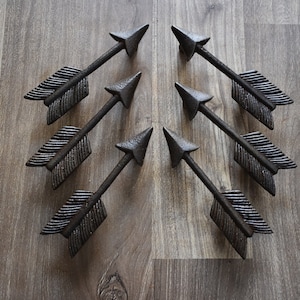 Könnte beinhalten: Acht rustikale, pfeilförmige Schrankgriffe in dunkelbrauner Ausführung. Die Griffe sind symmetrisch auf einer Holzoberfläche angeordnet.