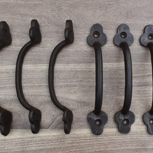Könnte beinhalten: Sechs schwarze Türgriffe aus Gusseisen mit geschwungenem Griffdesign. Die Griffe haben eine dekorative blütenartige Form an den Befestigungspunkten. Die Hardware wird auf einer hellbraunen Holzoberfläche gezeigt.