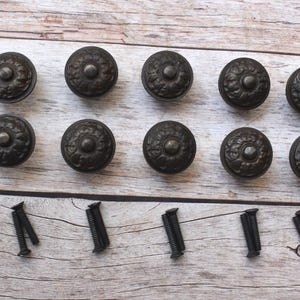 Puede incluir: Diez tiradores de armario de bronce antiguo con diseños florales decorativos dispuestos sobre una superficie de madera clara. Los tiradores son redondos con un detalle central en relieve. También se incluyen tornillos negros.
