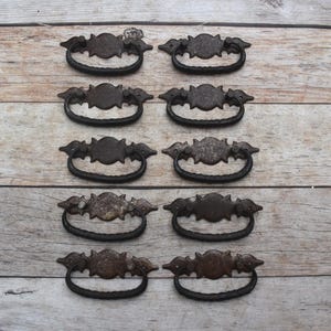 May include: A set of ten dark brown metal drawer pulls arranged in two vertical columns. Each pull features a central oval mounting plate with decorative pointed edges and a curved, textured drop handle attached to the base plate.