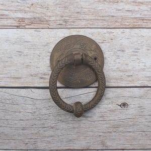 Peut inclure: Un heurtoir de porte circulaire en bronze doté d'un anneau texturé avec un motif de feuilles et d'une plaque décorative centrale. La plaque présente un motif floral en relief avec des détails de vigne complexes pour une porte.