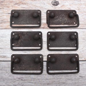 May include: A set of six rectangular metal drawer pulls with a dark, weathered, and rusted finish. Each pull features a flat backplate with two mounting holes on each side and a hinged, U-shaped handle attached by two rounded metal studs.