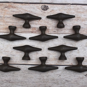Puede incluir: Nueve tiradores de armario de color bronce oscuro dispuestos sobre una superficie de madera clara. Cada tirador tiene un diseño en forma de T con una base redondeada y una parte superior plana en forma de ala. Los tiradores están espaciados uniformemente.