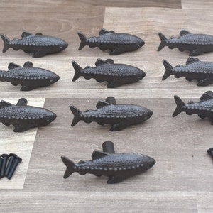 Könnte beinhalten: Zehn schwarze, gusseiserne, fischförmige Schubladenknöpfe mit strukturierter Oberfläche. Die Knöpfe sind in zwei Reihen zu fünf angeordnet.