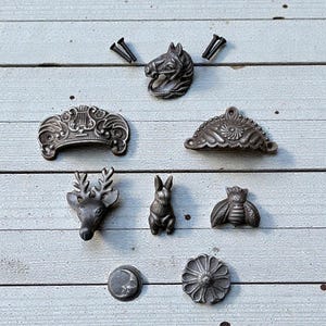 Puede incluir: Una colección de herrajes para gabinetes de estilo antiguo en varios diseños, incluyendo una cabeza de caballo, una cabeza de ciervo, un conejo, una abeja y tiradores decorativos. Los herrajes son de metal oscuro y están dispuestos sobre una superficie de madera clara.